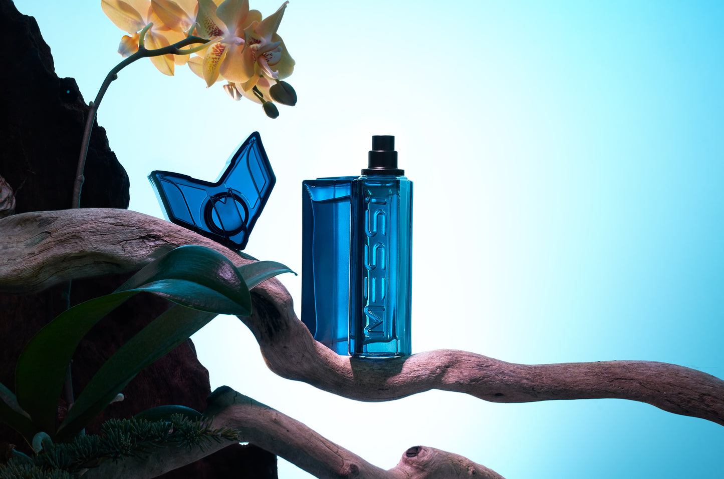 Messi Eau de Parfum bottle held by a branch with a yellow flower in the background, set against a bright blue backdrop.